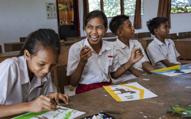 Three students in white and red uniforms sit at a long wooden desk in a bright classroom, engaged in an art activity. In the center, a girl laughs joyfully while holding a small paint tube.