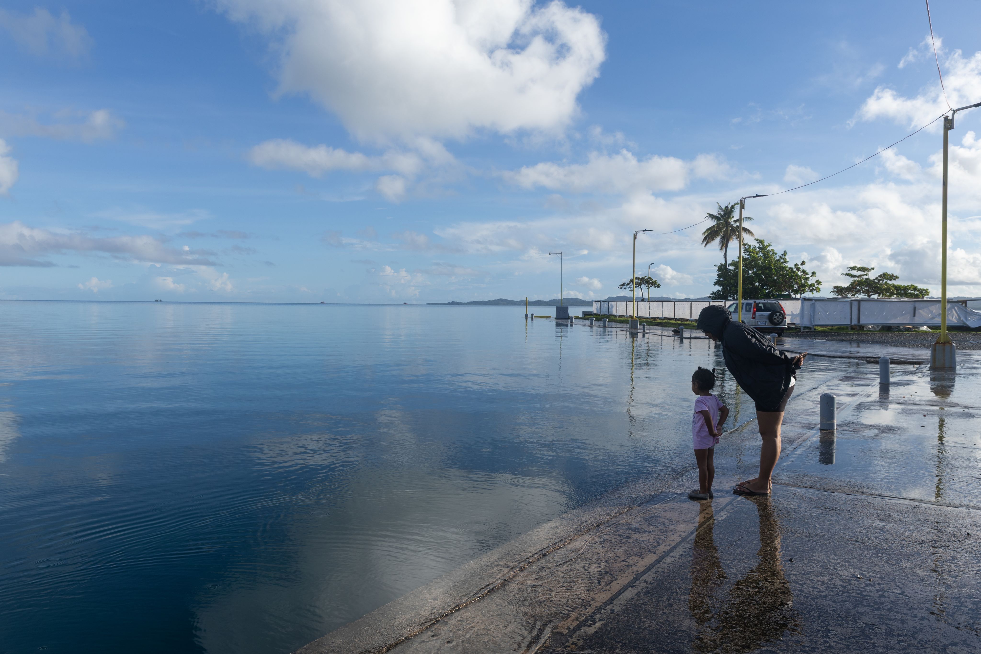 A woman and a small child stand on the edge of a dock. Water from the ocean is seen flooding the surface of the dock.